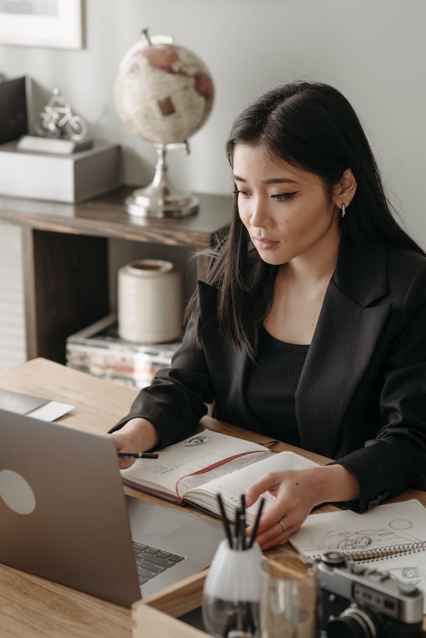 Asian woman in office attire focused on work at a desk with a laptop and notebook.